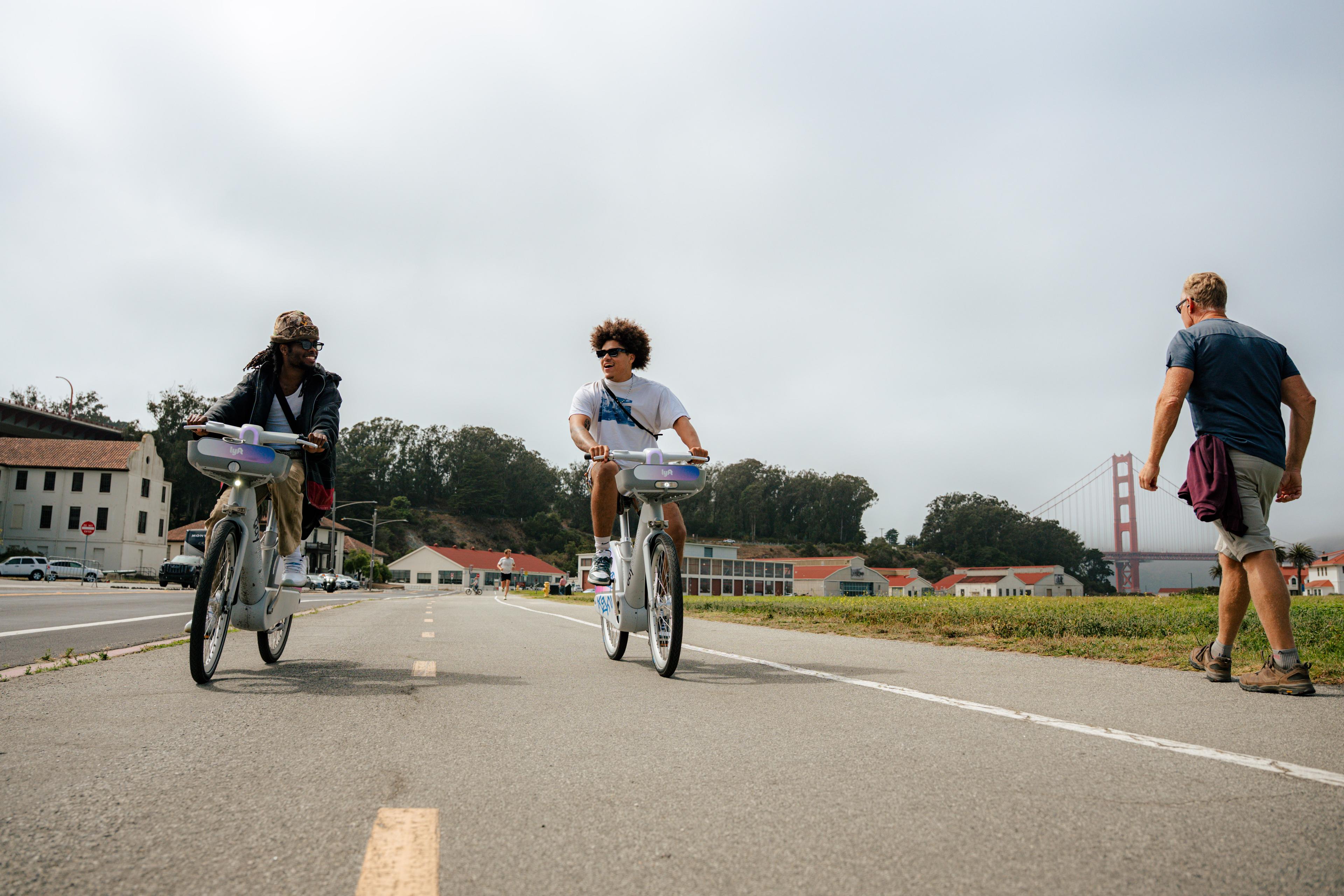 Two people biking next to each other with a man walking beside them with the golden gate bridge in the background,